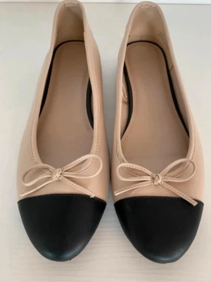 Beige ballerinaskor med svart tå - Snygga ballerinaskor i beige med svart tåparti och dekorativ rosett framtill. Skorna har rundad tå och är tillverkade i ett slätt material som ger en klassisk look. Perfekta för dig som gillar stilrena detaljer.