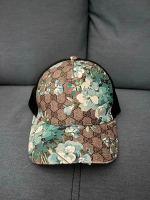 Gucci keps med blå blommor - Snygg keps från Gucci med klassiskt GG-mönster och blåa samt turkosa blommor över hela kepsen. Justerbar rem bak med grön-röd detalj. Materialet är en mix av canvas och mesh, vilket gör den både luftig och bekväm. Perfekt för dig som vill sticka ut med en unik accessoar.