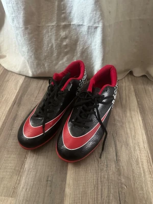 Svarta och röda fotbollsskor - Säljer ett par snygga fotbollsskor i svart och rött med vita detaljer. Skorna har snörning, låg profil och mönstrad häl i vitt. Insidan är röd och yttermaterialet är syntetiskt med struktur för bättre bollkontroll. Knappt använda. Går även att hämta i centrala Kungälv, om så önskas 