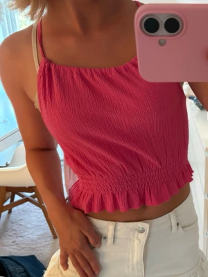 Rosa croppad linnetopp med smock - Säljer en rosa croppad linnetopp med smock och volangkant nertill. Toppen har tunna axelband som korsas i ryggen och ger en öppen rygg. Perfekt för sommarens varma dagar och enkel att styla med kjol eller jeans.