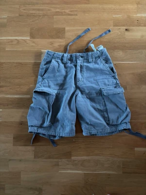 Blå cargoshorts med snörning - Säljer ett par blå cargoshorts med flera praktiska fickor och snörning i midjan och vid bensluten. Shortsen har en avslappnad passform och är perfekta för sommardagar. Tillverkade i bomull för en skön känsla.