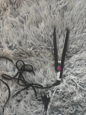Svart plattång med lila knapp - Säljer en svart plattång med vit och lila detalj. Plattången har sladd och vanlig stickkontakt, perfekt för att styla håret snabbt och enkelt. Smal och smidig design som gör den enkel att använda och ta med sig.
