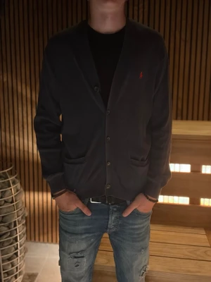 Mörkblå cardigan med knappar - Snygg mörkblå cardigan med knäppning framtill och två fickor. Klassisk design med V-ringning och diskret röd logga på bröstet. Perfekt att slänga över en t-shirt för en avslappnad men stilren look.