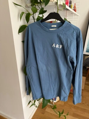 Blå långärmad tröja A&F - Blå långärmad tröja med rund hals och vit A&F-logga på bröstet. Tillverkad i mjukt bomullstyg och har en avslappnad passform. Perfekt för en casual look och enkel att matcha med jeans eller byxor.