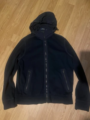 Svart vindjacka med huva - Snygg svart vindjacka med dragkedja framtill och två fickor med dragkedjor. Jackan har en justerbar huva och är tillverkad i ett lätt material som passar perfekt för blåsiga dagar. Enkel och stilren design som funkar till det mesta.