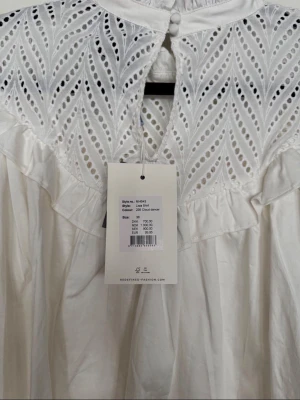 Vit blus med spetsdetaljer - storlek 38  - Supersöt vit blus med broderade spetsdetaljer och volang längs axlarna. Blusen har hög krage med knappar i nacken och långa ärmar med breda manschetter. Perfekt för dig som gillar romantisk och trendig stil.
