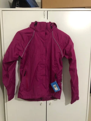 Rosa vindjacka med huva - Snygg rosa vindjacka med huva och dragkedja framtill. Jackan har två fickor med dragkedja och reflekterande detaljer längs sömmarna. Tillverkad i lätt och vindtätt material, perfekt för blåsiga dagar. Justerbar huva och ärmslut för extra komfort.