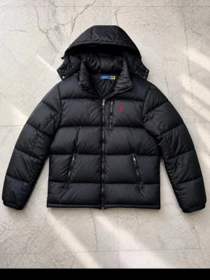 Svart pufferjacka Polo Ralph Lauren - Svart pufferjacka från Polo Ralph Lauren med huva och dragkedja framtill. Jackan har två dragkedjeförsedda fickor och en liten röd logga broderad på bröstet. Perfekt för kalla vinterdagar och riktigt skön att bära.