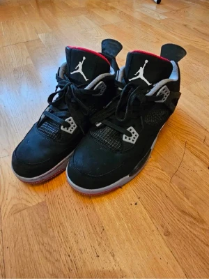 Svarta Air Jordan 4  - Säljer ett par svarta Air Jordan 4 sneakers med grå och röda detaljer. Skorna har meshpaneler, synlig Air-sula och Jumpman-logga på plösen. Ovandelen är i mocka och textil, och de har klassiska plastdetaljer vid snörningen. Priset kan diskuteras