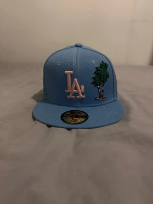 Ljusblå  keps med palmer - Säljer en ljusblå New Era 59FIFTY keps med broderad -logga i ljusrosa och två gröna palmer på framsidan. Klassisk flat brim och strukturerad passform. Perfekt för dig som vill sticka ut med en unik och färgglad keps.