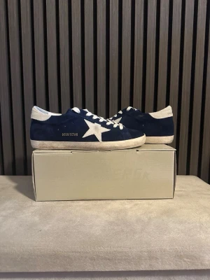 Mörkblå Golden Goose sneakers - Snygga mörkblå sneakers från Golden Goose med vit stjärndetalj på sidan, vita skosnören och vit sula. Skorna har en klassisk låg modell och är tillverkade i mocka med kontrasterande vita detaljer vid häl och tå. Perfekta för en trendig och avslappnad stil.