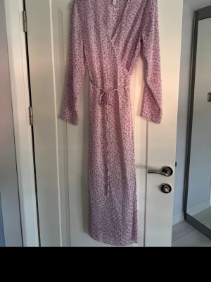 Ljuslila blommig omlottklänning H&M - Säljer en lång, ljuslila omlottklänning från H&M med blommigt mönster. Klänningen har långa ärmar, knytband i midjan och är tillverkad i ett lätt och luftigt material, perfekt för varmare dagar.