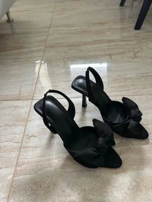 Svarta pumps med rosett, storlek 38 - Stilrena svarta pumps med öppen tå och bred klack. Skorna har en elegant rosett framtill och smala remmar runt hälen. Tillverkade i ett material som ser ut som satin, vilket ger en lyxig känsla. Perfekta för dig som vill ha en chic och trendig look.