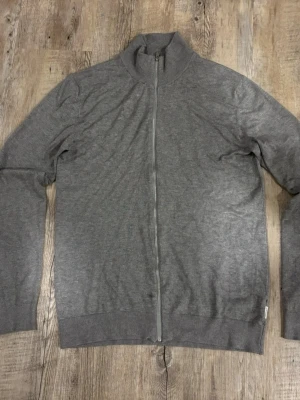 Grå ziptröja från Lindbergh - Stilren grå ziptröja från Lindbergh i slim fit-modell. Tröjan har hög krage, hel dragkedja framtill och ribbade muddar vid ärmslut och nederkant. Perfekt att slänga på sig över en t-shirt för en clean look.