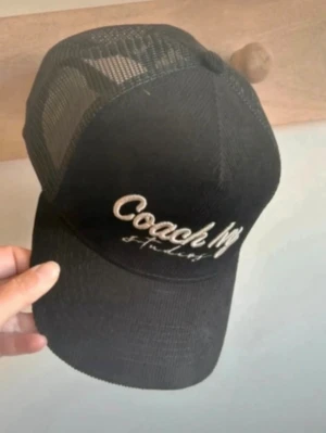 Svart keps Coach My Studios - Snygg svart keps med vit brodyr där det står 'Coach Ivy Studios' på framsidan. Kepsen har klassisk trucker-modell med mesh på sidorna och justerbar rem bak. Materialet framtill är ribbat tyg som ger en cool struktur.