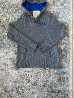 Grå hoodie från Hollister med blått foder - Snygg grå hoodie från Hollister med blått foder i huvan och blå logga på bröstet. Tröjan har dragsko i huvan, ribbade muddar och fickor framtill. Perfekt för en avslappnad och cool stil.