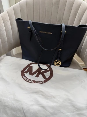 Mörkblå handväska Michael Kors - Snygg mörkblå handväska från Michael Kors i tåligt saffianoläder. Väskan har guldfärgade detaljer, dubbla axelremmar och ett dekorativt MK-hänge. Klassisk och rymlig modell med dragkedja upptill och stilren design.