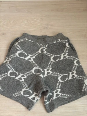 Grå stickade Cole buxton shorts - Säljer ett par grå stickade shorts med vitt CB-mönster över hela plagget. Shortsen har elastisk midja och två bakfickor. Perfekta för en avslappnad och trendig look.
