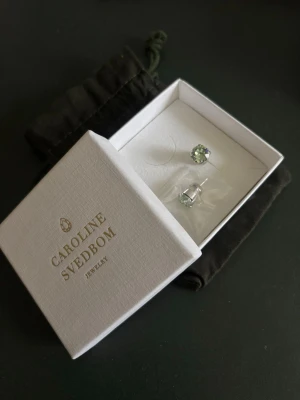 Caroline Svedbom örhängen  - Snygga örhängen från Caroline Svedbom med ljusgröna, gnistrande stenar i rund infattning. Levereras i original ask. Aldrig använda med plombering kvar! Orginalpris cirka 450kr.