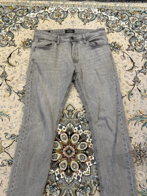 Ljusgrå tapered jeans - Säljer ett par ljusgrå jeans med tapered passform från Jack and Jones, storlek 30/30. och klassisk femficksdesign. Jeansen har bälteshällor, knappgylf och är tillverkade i ett mjukt denimtyg. Perfekta för en avslappnad och stilren look.