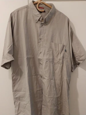Beige kortärmad skjorta med ficka - Stilren beige skjorta i bomull med korta ärmar och klassisk krage. Skjortan har en bröstficka och knäppning framtill. Perfekt för en avslappnad och enkel look.