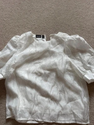 Vit blus med puffärm från Pieces - Säljer en vit blus från Pieces med puffiga korta ärmar och diskret blommönstrad struktur i tyget. Blusen har rund halsringning och är gjord i ett lätt, något glansigt material som ger en elegant känsla. Perfekt för att lyfta vilken outfit som helst.