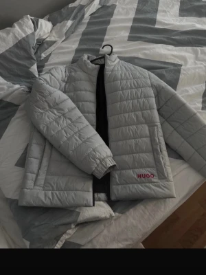 Ljusgrå dunjacka från HUGO - Snygg ljusgrå dunjacka från HUGO med rakt snitt och diskret quiltad design. Jackan har två sidofickor och HUGO-logga i rött framtill. Perfekt för kalla vinterdagar och ger en clean look.
