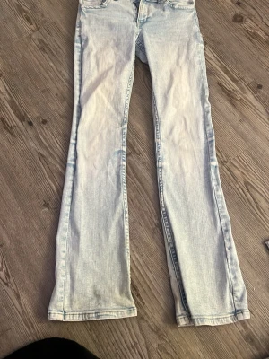 Ljusblå bootcut jeans - Säljer ett par ljusblå bootcut jeans med klassisk femficksmodell och dubbla knappar i midjan. Jeansen har en snygg tvättad look och raka, något utsvängda ben. Perfekta för dig som gillar retrostil och vill ha ett par bekväma jeans i bomull.