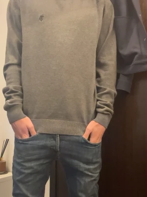 Grå basic  polo tröja - En stilren grå tröja med rund hals och diskret broderad logga på bröstet. Tröjan har långa ärmar, ribbade muddar och är gjord i ett mjukt material som passar perfekt till jeans. Enkel och klassisk design som funkar till det mesta.