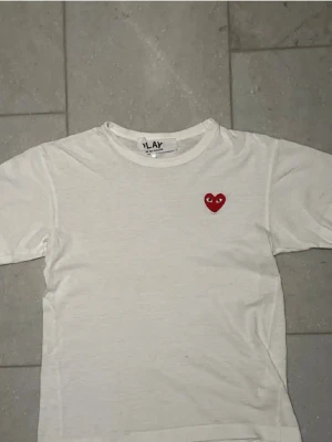 Vit t-shirt med rött hjärta - Vit t-shirt med ett litet rött hjärta med ögon broderat på bröstet. Klassisk rund hals och korta ärmar. Perfekt för dig som gillar enkel men snygg stil.