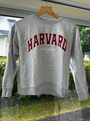 Grå Harvard sweatshirt - Säljer en grå sweatshirt med röd HARVARD-text och vit text under som säger UNIVERSITY BOSTON. Tröjan har rund halsringning, långa ärmar och ribbade muddar vid ärmslut och nederkant. Perfekt för dig som gillar college-stil. Storlek M.