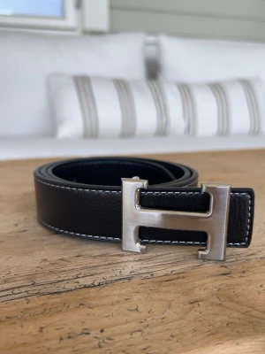 Herme’s bälte med silverfärgat H-spänne - Stilrent svart skärp med ett stort, silverfärgat H-format spänne. Skärpet har vita kontrastsömmar längs kanterna och är tillverkat i skinn. Perfekt accessoar för att lyfta din outfit med en klassisk touch. Bältet är aldrig använt.
