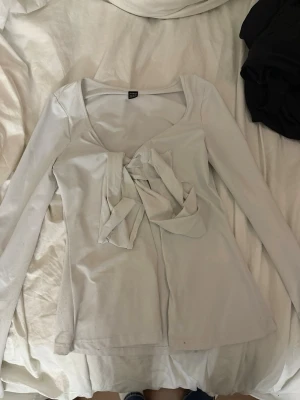 Vit långärmad topp med rosett SHEIN - Snygg vit långärmad topp från SHEIN med stor dekorativ rosett framtill. Toppen har en feminin v-ringning och är tillverkad i ett mjukt material som sitter bekvämt på kroppen. Perfekt för dig som vill ha en söt och trendig look.