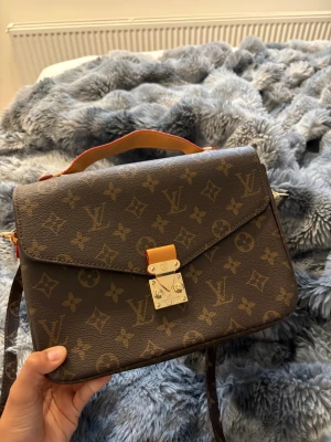 monogram handväska - Säljer en klassisk brun handväska från  med ikoniskt monogrammönster och guldfärgat lås. Väskan har ett handtag i ljust läder och axelrem. Perfekt för dig som vill ha en lyxig accessoar med tidlös design. Helt oanvänd, ej äkta