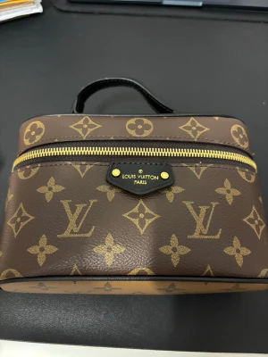 Monogram handväska - Säljer en klassisk brun handväska med det ikoniska monogrammönstret i beige och guldiga detaljer. Väskan har en guldfärgad dragkedja, svart handtag och detaljer i läder. Perfekt för dig som vill ha en lyxig accessoar med tidlös design.