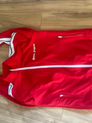 Röd track jacket från Palm Angels - Snygg röd track jacket från Palm Angels med vita detaljer och dragkedjor. Jackan har vit logga på bröstet, vita ränder längs ärmarna och hel dragkedja framtill. Perfekt för en sportig och trendig look. storlek s passar 170-180