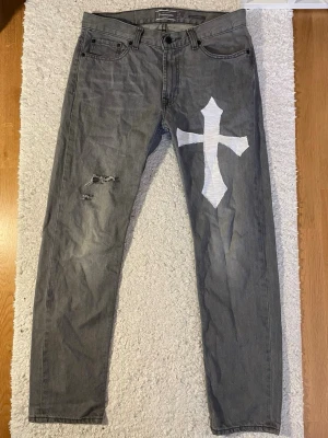 Grå jeans med vit korsdetalj - Säljer ett par grå jeans från Dressmann med en stor vit korsdetalj på höger ben och slitningar på vänster lår. Klassisk femficksmodell med knappgylf och raka ben. Materialet är denim i bomull. Perfekta för dig som vill sticka ut med din stil. L31 W30