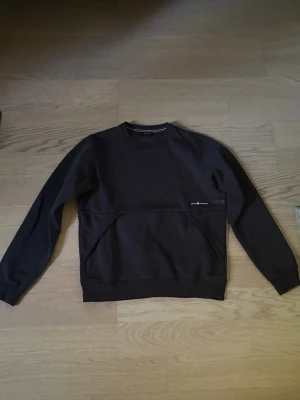 Svart sweatshirt från Sail Racing - Säljer en mörkbrun sweatshirt från Sail Racing med rund halsringning och diskret logga på bröstet. Tröjan har långa ärmar och en praktisk ficka framtill. Perfekt för en avslappnad och stilren look.