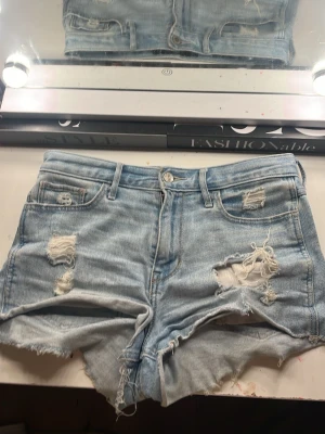 Ljusblå slitna jeansshorts - Säljer ett par ljusblå jeansshorts med slitna detaljer och fransig kant. Shortsen har fem fickor, bälteshällor och klassisk knappgylf. Perfekta för varma dagar och ger en avslappnad vibe.