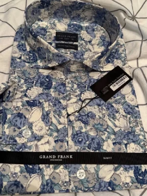 Blommig skjorta från Grand Frank - Säljer en slim fit skjorta från Grand Frank med blått och vitt blommönster. Skjortan har klassisk krage, knappar framtill och bröstficka. Perfekt för dig som vill sticka ut med en färgglad och stilren look. Aldrig använd.