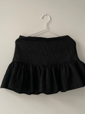Svart volangkjol från Zara - Kort svart kjol från Zara med volang nedtill och lätt struktur i tyget. Kjolen har en hög midja och en feminin siluett som ger en trendig look. Perfekt att matcha med en enkel topp eller stickad tröja.