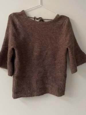 Brun stickad topp med öppen rygg - Mysig brun stickad topp med trekvartsärmar och bred ribbad kant nedtill. Toppen har en öppen rygg med knytband upptill för en snygg detalj. Perfekt att styla med jeans eller kjol. Materialet känns mjukt och skönt mot huden.