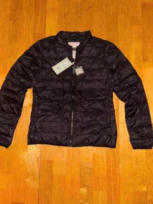 Svart lättvikt pufferjacka - Snygg svart pufferjacka med quiltad design och dragkedja framtill. Jackan har en minimalistisk look utan huva och är tillverkad i ett glansigt, lätt material som håller värmen. Perfekt för kalla vinterdagar och enkel att matcha med olika outfits.