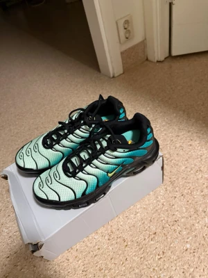 Nike Air Max Plus turkos/svarta sneakers - Säljer ett par Nike Air Max Plus sneakers i turkos, svart och vitt med gula detaljer. Skorna har en vågig design på ovandelen, svarta snören och en robust sula med synliga luftbubblor. Perfekta för dig som gillar streetwear och vill sticka ut.