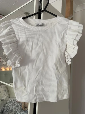 Vit ribbad topp med volangärmar Zara - Supersöt vit ribbad topp från Zara med bred rund halsringning och stora volangärmar som ger en extra touch till outfiten. Perfekt att styla med jeans eller kjol för en trendig look. Materialet känns mjukt och bekvämt. Den har någon fläck men har inte testat tvätta den