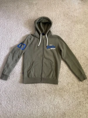 Superdry Zip Hoodie - Grön zip hoodie från Superdry med blå och vita detaljer. Dragkedja framtill, huva och två fickor.  I mycket fint skick och säljs då den inte används längre.  📦 Snabb leverans! 💬 Hör gärna av dig vid frågor eller om du vill ha fler bilder/mått!