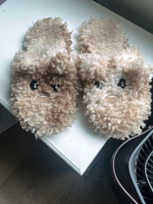 Beiga fluffiga tofflor med ansikte - Säljer ett par supermysiga tofflor i beige och ljusbrun färg med lockigt, fluffigt material. Tofflorna har ett gulligt ansikte broderat framtill och är öppna baktill för enkel påtagning. Perfekta för att hålla fötterna varma hemma. Pris diskuteras