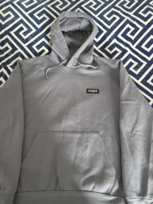 Grå hoodie från McKenzie - Säljer en stilren grå hoodie från McKenzie med huva och dragsko. Hoodien har en klassisk känguruficka framtill och en liten svart logotyp på bröstet. Perfekt för en avslappnad och casual look.