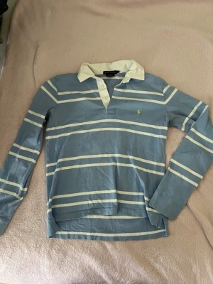 Blåvit randig långärmad tröja Ralph Lauren - Säljer en blå och vit randig långärmad skjorta från Ralph Lauren i 100% bomull. Skjortan har klassisk krage, knappar vid halsen och en broderad logga på bröstet. Perfekt för en avslappnad men ändå stilren look.