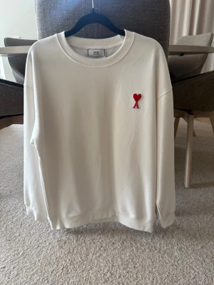 Ami sweatshirt  - Säljer en stilren vit sweatshirt från Ami med deras ikoniska röda hjärta och A på bröstet. Tröjan har rund halsringning, ribbade muddar och är tillverkad i mjukt bomullsmaterial. Perfekt för en clean och trendig look.